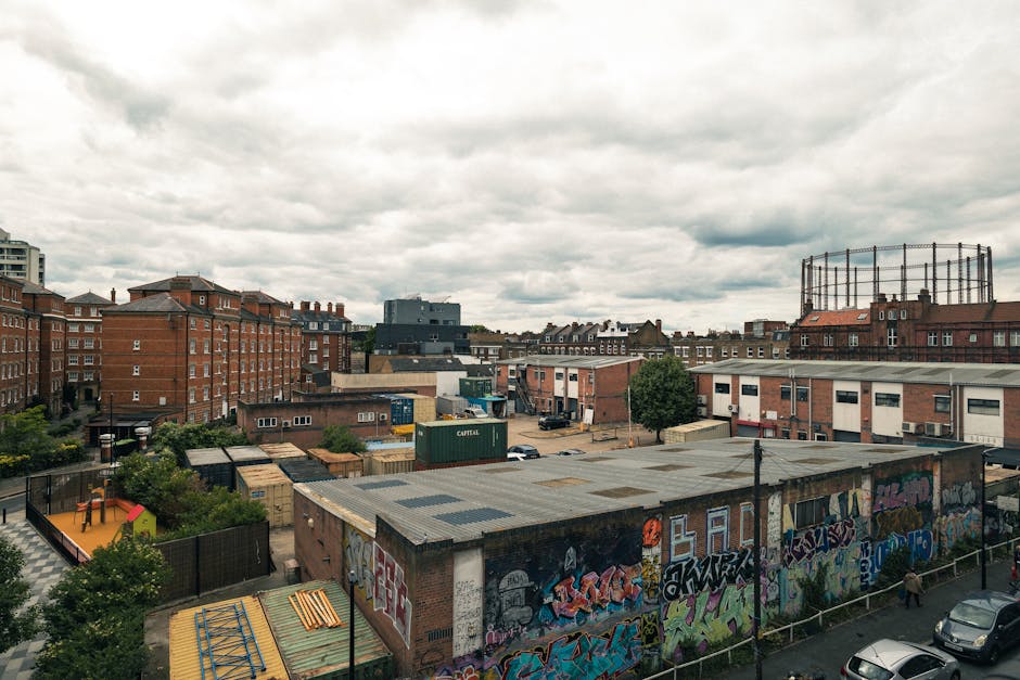 A daytime urban scene shows a variety of buildings, including red-brick residential flats on the left and mid-rise commercial structures on the right, set against a cloudy sky. In the foreground, there is a large, flat-roofed building covered with colorful graffiti art. Behind this building, a fenced playground with a small slide and climbing frame is visible, surrounded by green trees and bushes. Several vehicles are parked in the open area and along the street, with shipping containers stacked nearby, indicating an industrial or storage zone. A round, metal water tank on a high platform is positioned on the far right, while other warehouse-like buildings with flat roofs make up the background, creating a mix of residential, commercial, and industrial environments. The scene is lit with diffuse natural light typical of an overcast day, highlighting the textured surfaces of the rooftops, brickwork, and graffiti, reflecting the typical urban landscape associated with alternative waste handling or independent rubbish removal services such as those offered by Rubbish Collection Hackney.