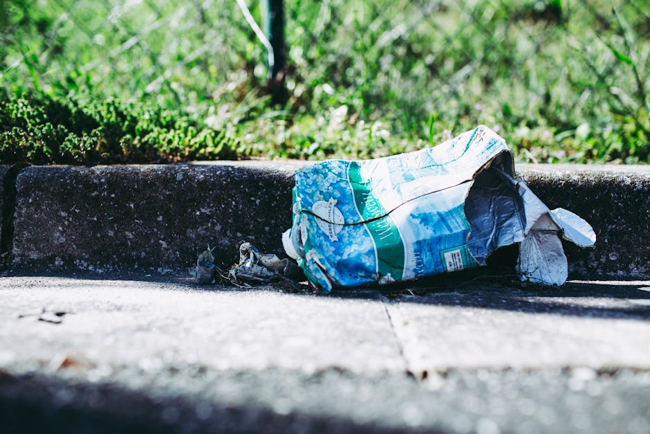 A crumpled plastic bag made of thin, glossy material with a predominantly blue and white design, lying discarded on a concrete sidewalk near a raised curb. The bag appears partially torn, with some plastic debris spilled out, and is positioned close to a patch of uneven pavement with visible cracks and small debris scattered around. Behind the curb, there is a lush, green grassy area with dense blades of grass and some small plants. The scene is illuminated by natural daylight, creating shadows on the ground and highlighting the textures of the plastic bag, pavement, and grass. This image exemplifies the presence of litter in an outdoor urban environment, illustrating the importance of proper waste disposal, which is a key aspect of private rubbish collection services like those offered by Rubbish Collection Hackney and other rubbish removal specialists.
