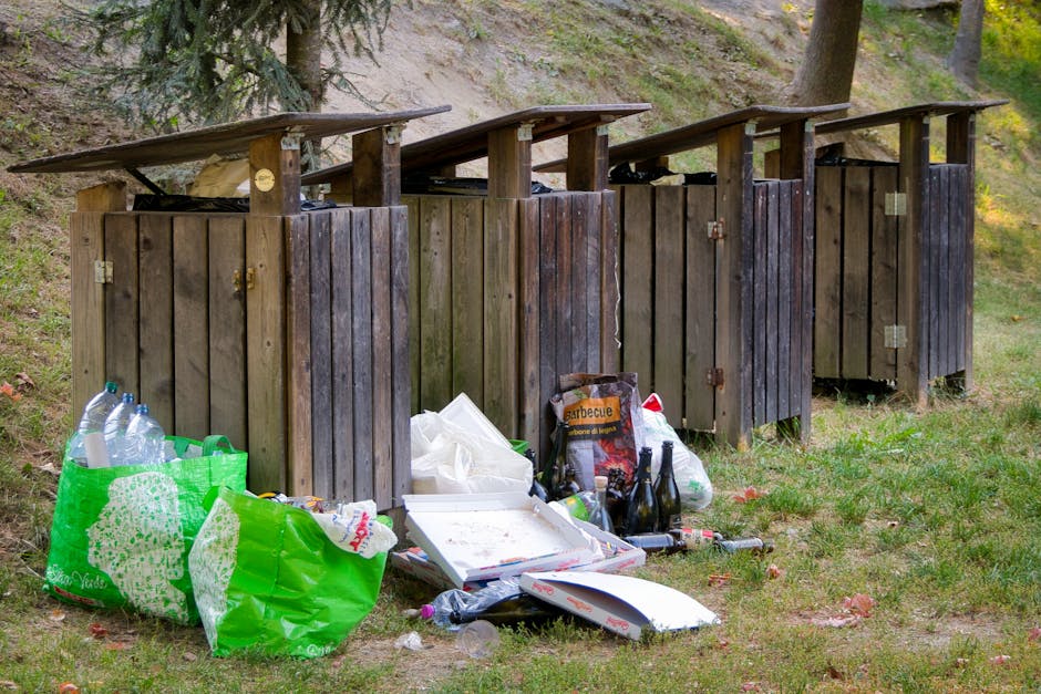 Four wooden waste bins with slanted roofs made of weathered, dark-stained planks are positioned on a grassy area near a sloped hillside with trees in the background. In front of the bins, there is a collection of household rubbish including a green reusable shopping bag filled with plastic bottles, a white pizza box, a black plastic bag, and several glass bottles, some of which are dark brown or green. The waste is scattered on the ground, which appears slightly muddy with patches of grass and scattered fallen leaves. The scene is outdoors, illuminated by natural light, and the overall impression is of an informal or inappropriate disposal of waste, possibly reflecting issues with rubbish collection or private waste handling services such as those offered by Rubbish Collection Hackney. The arrangement of the bins and scattered rubbish suggests an area designated for waste but not properly maintained or cleared regularly.