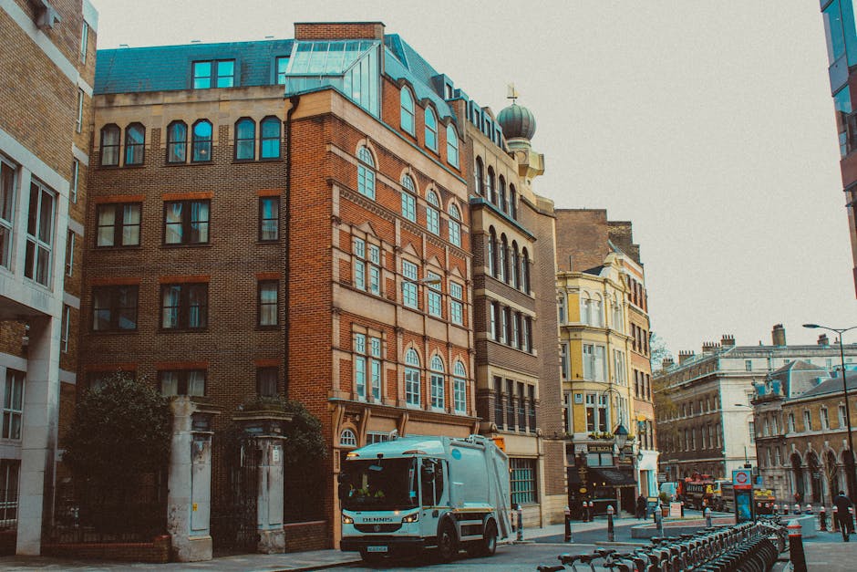 A street scene featuring multi-storey buildings with a mix of brick and ornate architectural details, including arched windows and decorative facades. In the foreground, a small rubbish collection vehicle with a light blue body and black wheels is parked on the street, partially blocking the view of the pavement. The vehicle is positioned near a gated entrance with stone pillars, and the street is lined with metal bike racks and bollards. The surrounding environment includes older, historic-style buildings with turreted roofs and a dome visible on one structure, alongside modern establishments. The lighting suggests a cloudy day, with diffuse natural light illuminating the scene. This setting exemplifies urban waste management in an inner-city area, where independent collection services such as those provided by Rubbish Collection Hackney may operate alongside municipal services, offering alternative waste handling solutions in a densely built environment.