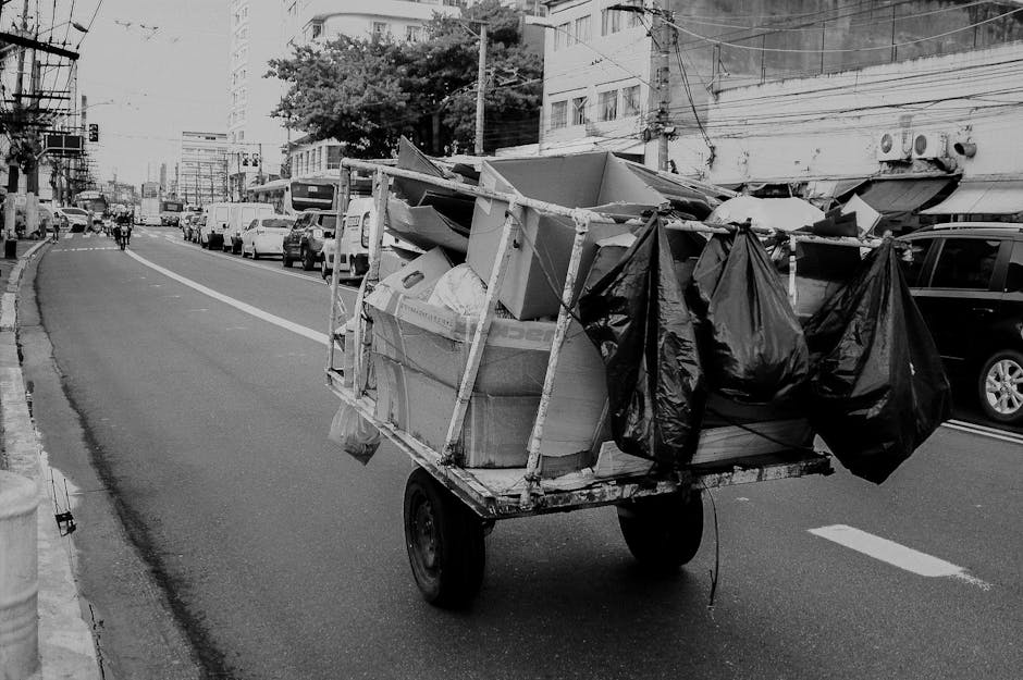 A small, manually propelled cart is loaded with various types of waste materials, including large cardboard boxes and black plastic garbage bags, positioned on a city street. The cart has a metal frame and is placed on two large wheels, with the cardboard boxes stacked at the front and secured with wooden supports, some of which appear tilted or slightly dislodged. The black plastic bags are hanging from the sides of the cart, possibly filled with additional waste. The street is lined with parked vehicles, including cars and vans, and features a mix of commercial and residential buildings in the background, some with visible air conditioning units and electrical wiring. The environment appears urban with a slight downward slope, and the street surface shows typical asphalt pavement with painted lane markings. The scene suggests an instance of independent waste collection, possibly related to non-contracted rubbish removal services, illustrating a form of private on-site rubbish handling in an urban location related to Hackney's rubbish management activities.