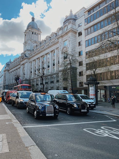 A street scene in an urban area featuring multiple black taxi cabs parked along the side of the road in front of a historic white stone building with classical architectural elements, including columns and ornate details, topped with a dome. Behind the taxis, there are several other vehicles, including a red double-decker bus and a white van, indicating typical city traffic. On the right, a bus shelter with a digital timetable is situated next to sidewalk pedestrians, some walking and others waiting. The building has large windows reflecting the blue sky with scattered clouds, and leafless trees line the street, suggesting late autumn or winter. The scene is captured during daylight with natural lighting, highlighting the textures of the stone façade and the various vehicle finishes, emphasizing an active city environment that could relate to alternative or private waste collection servicing in busy commercial districts.