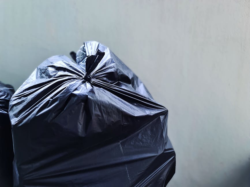 A small, manually propelled cart is loaded with various types of waste materials, including large cardboard boxes and black plastic garbage bags, positioned on a city street. The cart has a metal frame and is placed on two large wheels, with the cardboard boxes stacked at the front and secured with wooden supports, some of which appear tilted or slightly dislodged. The black plastic bags are hanging from the sides of the cart, possibly filled with additional waste. The street is lined with parked vehicles, including cars and vans, and features a mix of commercial and residential buildings in the background, some with visible air conditioning units and electrical wiring. The environment appears urban with a slight downward slope, and the street surface shows typical asphalt pavement with painted lane markings. The scene suggests an instance of independent waste collection, possibly related to non-contracted rubbish removal services, illustrating a form of private on-site rubbish handling in an urban location related to Hackney's rubbish management activities.
