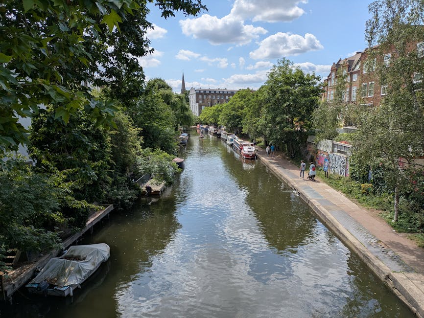 An aerial view of a narrow urban river lined with lush green trees on both sides, with several boats moored along the quay on the left bank, some covered with protective tarps. The right side features a pedestrian pathway where a few people are walking, adjacent to a row of red-brick residential buildings with white-framed windows. Cars are parked along the street parallel to the pathway, while some graffiti is visible on a fence near the trees. In the background, a church spire and additional buildings rise above the treetops under a partly cloudy sky with soft, natural daylight. The scene captures a peaceful, city-centre environment where waterway and urban life intersect, emphasizing the importance of managing local waste and refuse collection in residential areas, similar to services provided by Rubbish Collection Hackney for efficient rubbish removal and on-site clearance.