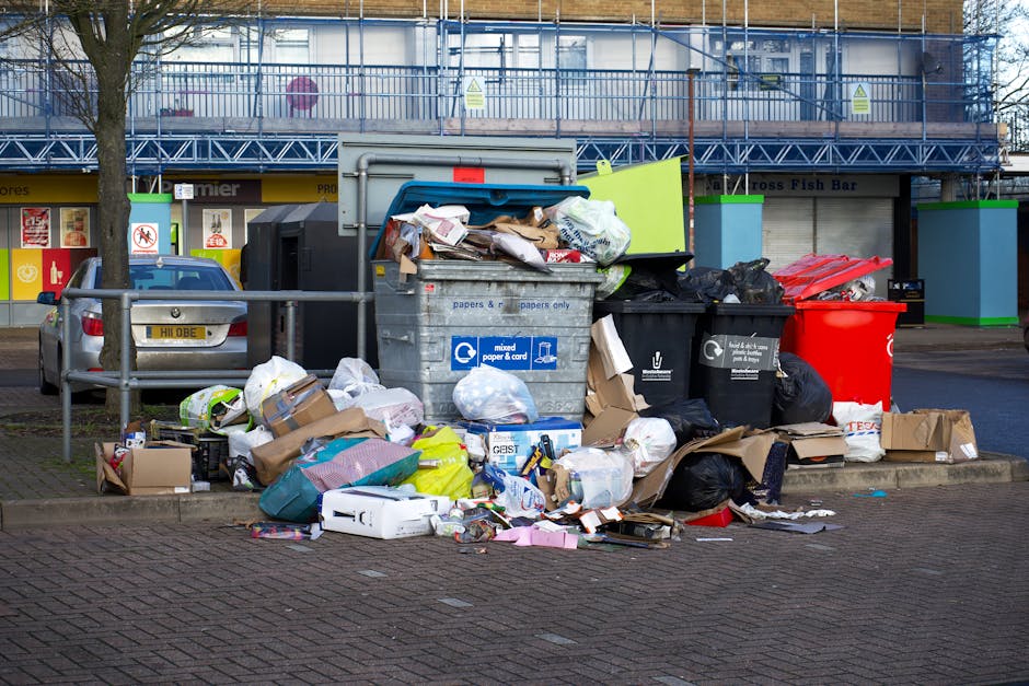 A large outdoor recycling and waste collection point situated on a paved urban area, featuring multiple open-top and closed waste bins. The central bin is a grey mixed paper and card recycling container, overflowing with crumpled paper, cardboard boxes, and various paper waste. To its right, there are black and black-and-white black bin bags filled with domestic waste, partially scattered on the ground, along with flattened cardboard boxes and discarded packaging materials. On the far right, a red waste bin is visible. Behind these, a low metal railing separates the waste area from parked cars, including a silver hatchback vehicle positioned towards the left of the scene. In the background, there is a commercial building with large storefront windows, displaying advertisements, and a shop sign partially visible. Above, there is a construction scaffold covering the upper facade of the building, with blue protective netting. The environment appears to be a busy urban setting where improper or overflow waste disposal is evident, highlighting potential challenges with private rubbish collection or disposal services managed by Rubbish Collection Hackney in local residential or commercial estates.