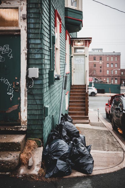 A residential building with green wooden cladding and red trim features a set of wooden stairs leading to an upper door with a small overhang. Several black garbage bags are piled against the building's lower wall, situated on a concrete sidewalk near a paved driveway. To the left, a doorway with a graffiti-covered metal door and concrete steps is visible, with patches of moss or dirt on the step edges. An electrical box with connected wiring is mounted on the building's exterior wall. In the background, parked cars and an urban landscape with brick buildings and power lines can be seen under overcast lighting, suggesting a typical street scene where private waste collection or alternative rubbish disposal methods may be employed by local residents or service providers like Rubbish Collection Hackney. This scene emphasizes the importance of proper rubbish storage and disposal in densely built environments, relating to the broader context of rubbish removal services for Hackney estates and alternative waste handling options.