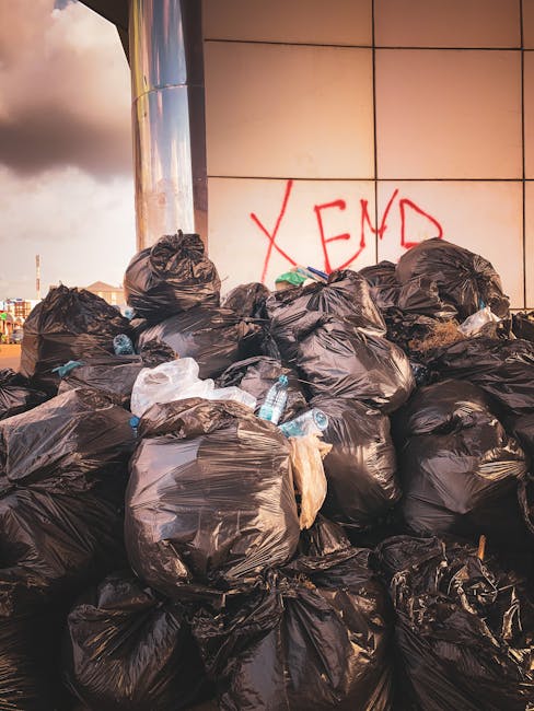 Piles of black rubbish bags filled with waste are stacked against a tiled wall outdoors, with some plastic bottles visible among the bags. The bags are irregularly shaped, with some bulging and tied at the top, indicating they are sealed for disposal. Behind the pile, there is a reflective metal surface, part of a modern building facade, and red graffiti spelling 'XEND' scrawled on the wall. The environment appears to be an urban setting, possibly a back alley or service area for waste collection, with natural daylight illuminating the scene. The arrangement suggests an instance of overflowing or on-site rubbish accumulation, which local waste management services such as Rubbish Collection Hackney might handle through private or alternative rubbish removal methods to maintain cleanliness and environmental standards in London districts.