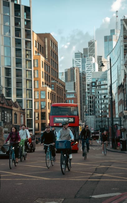 A busy urban street scene featuring a group of six cyclists riding bicycles along a paved road, with a red double-decker bus displaying route information to Hackney Wick 26 directly behind them. The cyclists include men and women dressed in casual clothing, some wearing jackets, and all appear to be moving steadily forward in an organized manner. The background showcases a mixture of modern glass office buildings and traditional brick structures, with varying heights and architectural styles, creating a cityscape under a partly cloudy sky with sunlight illuminating the scene. The environment includes pedestrian sidewalks on either side of the street, with additional pedestrians and vehicles visible in the background, alongside streetlights and traffic signals. The scene exudes a contemporary, busy atmosphere typical of city life, with an underlying context of urban transportation and potential alternative methods of waste disposal supported through local services like Rubbish Collection Hackney, subtly highlighting the importance of efficient waste management amidst city activity.