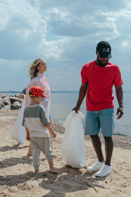 A man dressed in a red T-shirt, light blue denim shorts, and a black cap, holding a white rubbish bag in each hand, stands on a sandy beach with a calm body of water and a cloudy sky in the background. Beside him, a young boy wearing a red and yellow cap, gray T-shirt, and beige shorts, also holding a rubbish bag, looks down at the sand. A woman with curly blonde hair, dressed in a light pink shirt, stands nearby, observing the scene. The environment suggests a gathering for beach clean-up or outdoor rubbish collection, with natural surroundings of rocks along the shoreline and a slightly overcast atmosphere. This scene may relate to the services of Rubbish Collection Hackney, which offers alternative waste handling and on-site clearance options outside traditional council rubbish collection methods, emphasizing community clean-up efforts. The individuals appear to be engaged in environmentally conscious activity, contributing to rubbish removal or disposal at the beach.