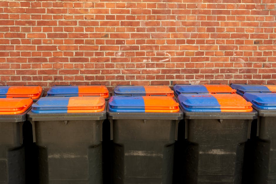 A row of six large, black plastic waste bins lined up on a paved surface against a red brick wall. Each bin has a partially visible, brightly coloured lid with a combination of blue, orange, and yellow sections. The lids are closed, and the bins are evenly spaced with no visible handles or labels. The environment appears to be outdoors, possibly near a building or a back alley, with natural daylight illuminating the scene. The image suggests a collection of refuse or waste awaiting disposal, fitting within the context of rubbish removal services like those provided by Rubbish Collection Hackney, highlighting the importance of organized waste management and alternative disposal options such as private collection or on-site clearance.